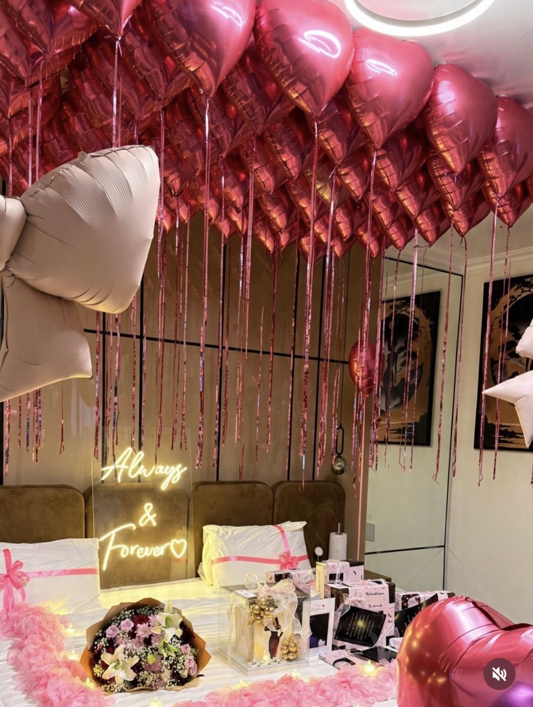 Pink heart balloon ceiling installation above a bed styled with a neon sign and gift boxes