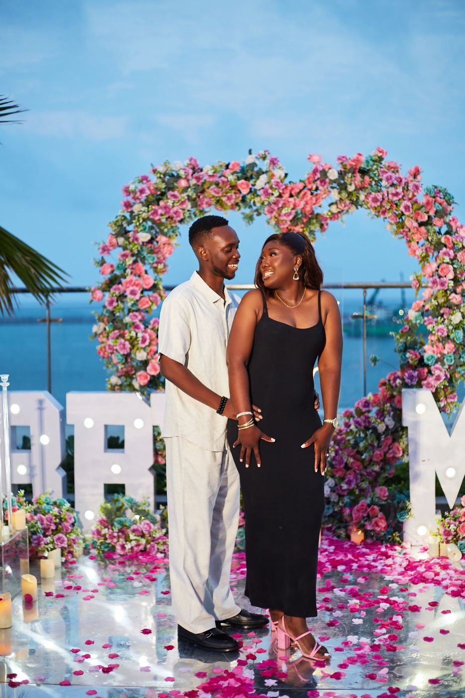 Couple smiling in front of a floral heart arch with a rooftop ocean view at dusk