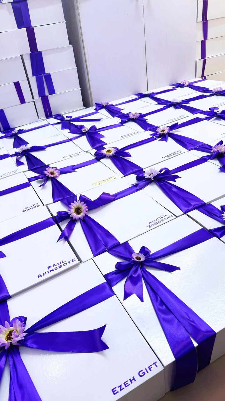 Rows of white gift boxes tied with purple ribbon and gerbera daisies, each labelled with a name
