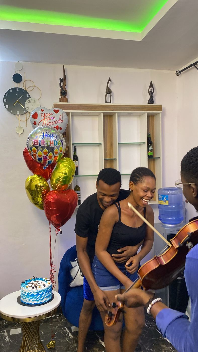 Couple laughing as a violinist serenades them beside a birthday cake and balloons