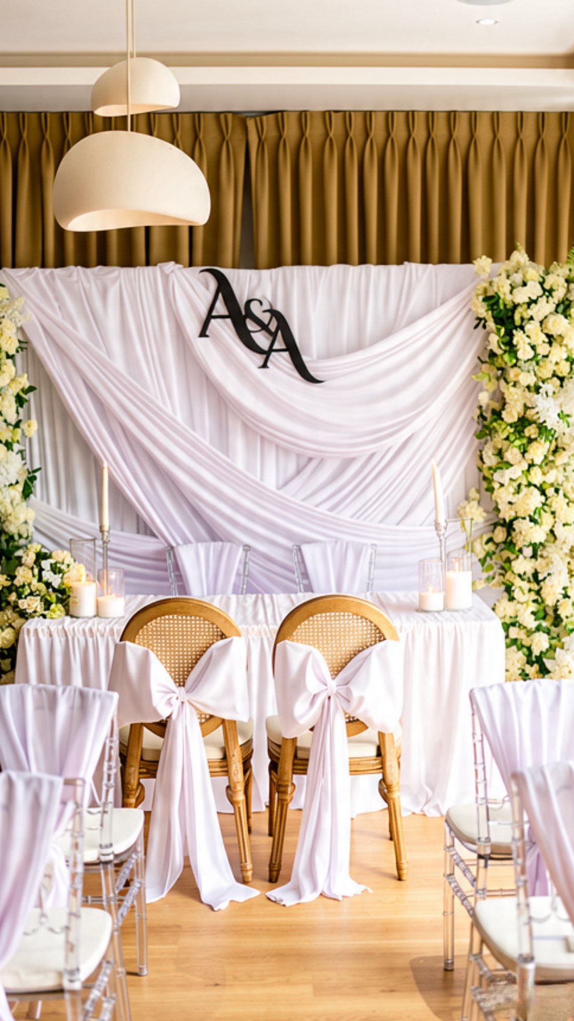 Draped white backdrop with an A&A monogram, floral pillars and chairs tied with white bows at a wedding ceremony